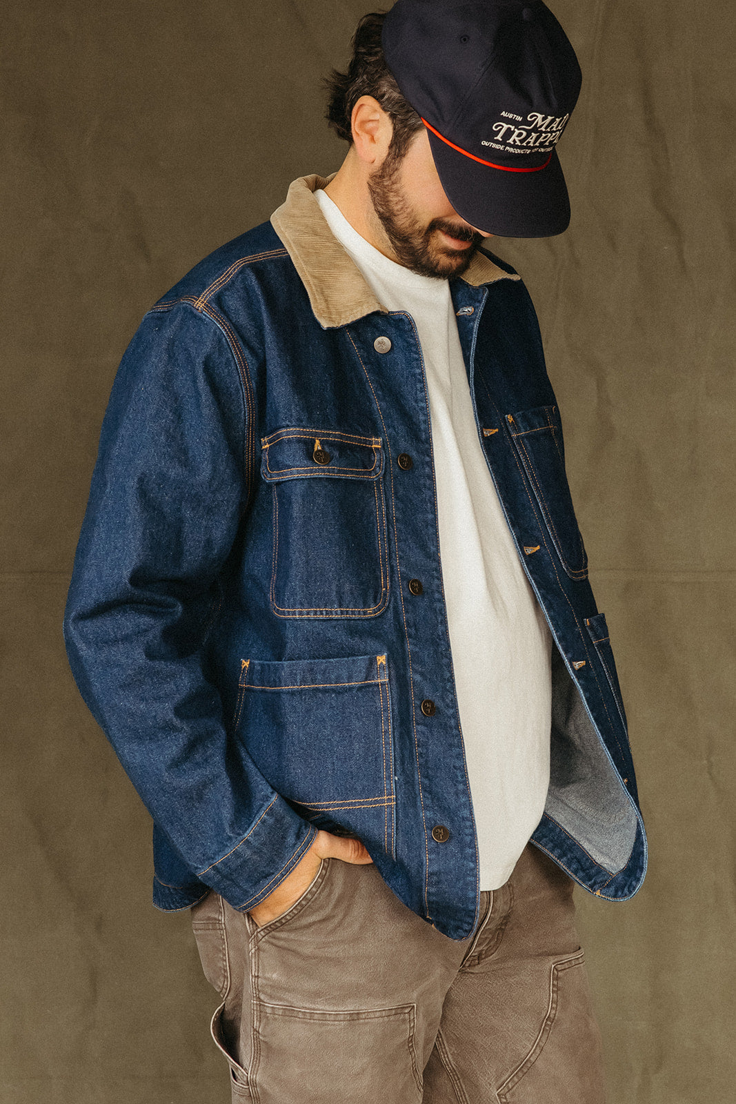 Man wearing a blue Mad Trapper Denim Field Coat white shirt, and brown pants against a brown background