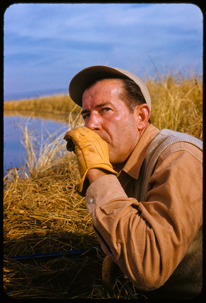 Mad Trapper image showing man duck calling next to body of water. 